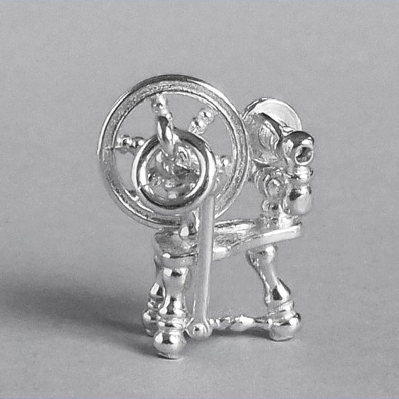 May include: A silver spinning wheel charm with a detailed design. The wheel is turned by a small handle and has a spindle for winding yarn.