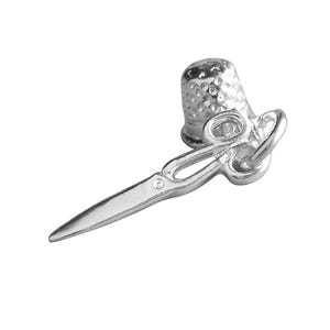 May include: A silver charm featuring a thimble and small scissors, likely for sewing or crafting. The thimble has a textured surface, and the scissors have a pointed tip. The charm is designed with a loop for attaching to a bracelet or necklace.