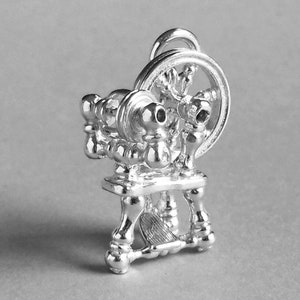 May include: A silver charm in the shape of a spinning wheel. The wheel is detailed with spokes and a handle. The charm is a traditional symbol of craftsmanship and creativity.