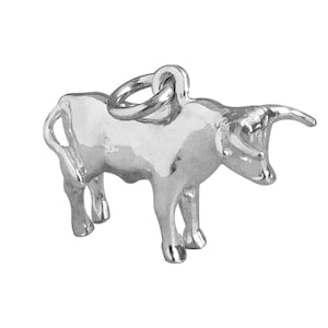 May include: Silver charm in the shape of a bull. The bull is standing with its head turned to the side and its horns pointing forward.