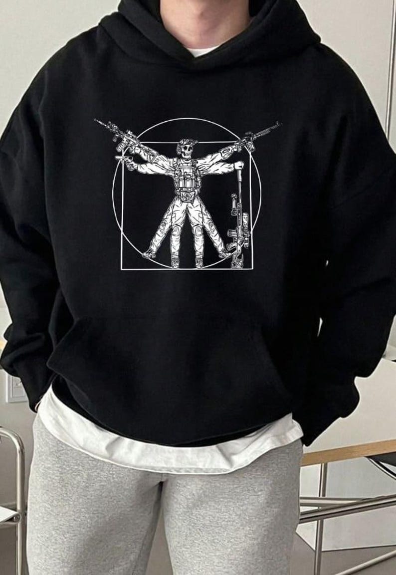 Peut inclure: Sweat &agrave; capuche noir avec un motif blanc repr&eacute;sentant une figure tenant des fusils, dans un cercle et un carr&eacute;. Le design rappelle l'Homme de Vitruve de Da Vinci. Le sweat &agrave; capuche a une capuche et un sous-v&ecirc;tement blanc est visible.