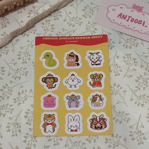 Può includere: Un foglio di adesivi con 12 diversi animali dello zodiaco cinese. Ogni animale è illustrato in uno stile cartone animato carino con uno sfondo giallo. Il foglio di adesivi è etichettato "Chinese Zodiacs Sticker Sheet by Anjoobi."
