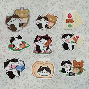 May include: A collection of colorful stickers featuring cartoon cats and bears. The stickers depict the characters with various food items, including boba tea, cake, and dango. One sticker has the text "Stay Hydrated". The stickers are on a floral background.