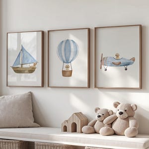 Può includere: Tre stampe incorniciate ad acquerello raffiguranti una barca a vela, una mongolfiera e un aereo, tutti con una combinazione di colori azzurro chiaro e marrone. Le stampe sono esposte sopra una panca con un cuscino, una casetta di legno e orsacchiotti.