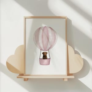 Könnte beinhalten: Ein gerahmter Aquarelldruck eines Heißluftballons mit rosa und weißen Streifen. Ein brauner Hund befindet sich im Korb. Das Kunstwerk wird auf einem Holzregal mit Wolkenmotiv ausgestellt.