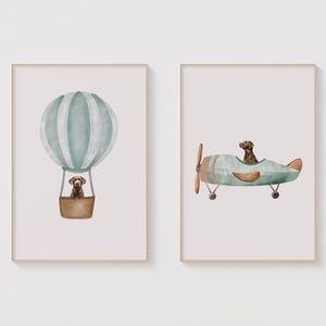 Könnte beinhalten: Zwei gerahmte Drucke mit einer Aquarell-Illustration eines Hundes in einem Heißluftballon und eines Hundes in einem Flugzeug. Der Heißluftballon ist grün und weiß und das Flugzeug ist grün und braun.