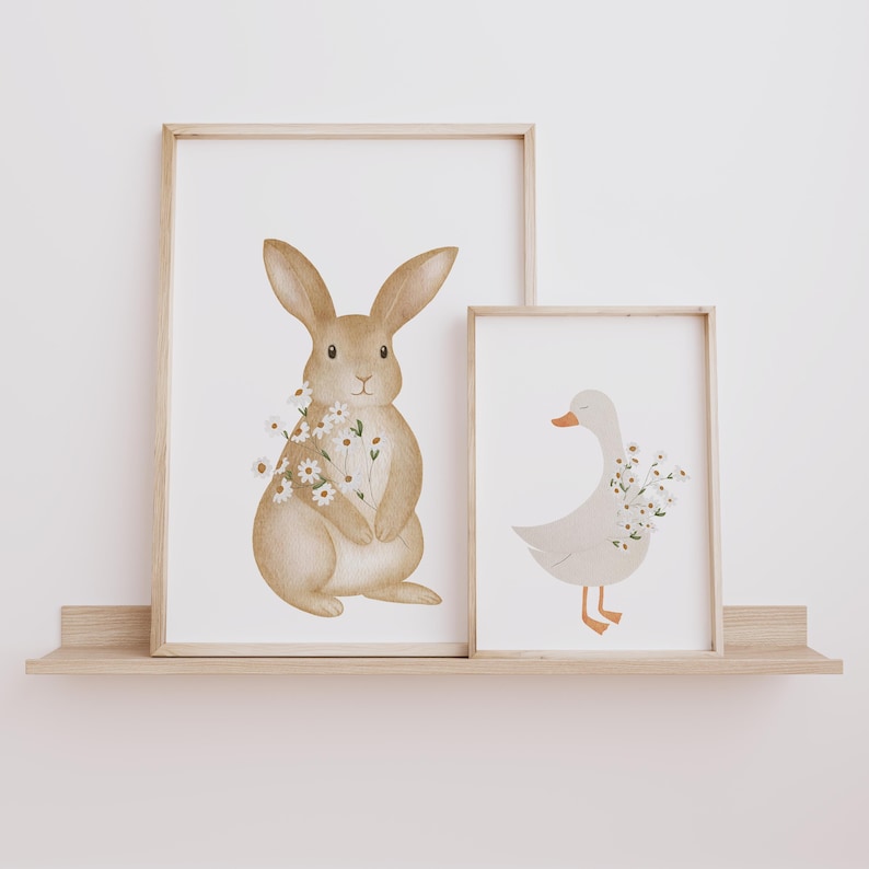 Puede incluir: Dos l&aacute;minas enmarcadas con ilustraciones en acuarela de un conejo y un ganso. El conejo es marr&oacute;n claro y sostiene flores. El ganso es blanco con pico y patas naranjas, tambi&eacute;n con flores. Ambas en marcos de madera clara.