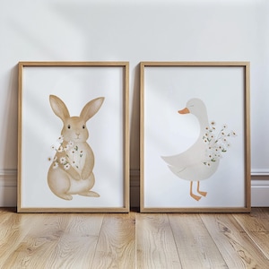 Puede incluir: Dos l&aacute;minas enmarcadas con ilustraciones en acuarela de un conejo y un ganso. El conejo es de color marr&oacute;n claro con un ramo de flores blancas. El ganso es blanco con pico naranja y flores blancas.