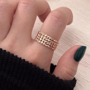 May include: A rose gold ring with a woven design is worn on a finger. The ring has a textured surface and a wide band. The person's fingernails are painted a dark green color. The background is a light beige color.