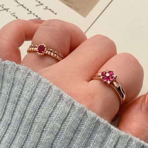 May include: Two gold rings are displayed on a hand. One ring features a small, oval, red gemstone. The other ring has a flower design with red gemstones and small white stones. The rings are on a light blue sweater.