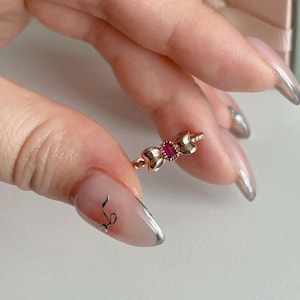May include: A rose gold ring featuring a bow design with a small, rectangular, red gemstone at the center. The ring is held up by a person's fingers, showcasing its delicate details and elegant style. The ring is a piece of jewelry.