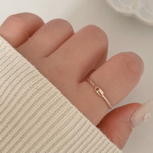 May include: A delicate gold ring with a rectangular center detail and a thin band. The ring is shown on a finger, with a white sweater in the background. The ring's design is minimalist and elegant.