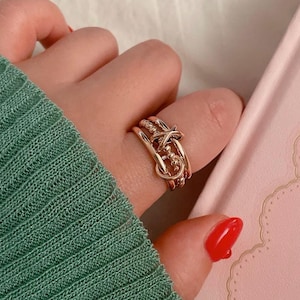 May include: A gold-toned ring with a knot design. The ring is worn on a finger with red nail polish.
