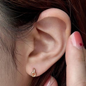 May include: A close-up of a gold-colored stud earring in the shape of a knot. The earring is in a person's ear. The person's hand is touching their ear. The person has pink nail polish with red heart designs.