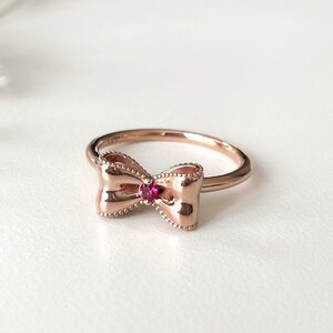 May include: A rose gold ring with a bow-shaped design and a small red gemstone in the center.