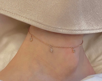 May include: A delicate gold anklet with three small charms hanging from it. Each charm is a diamond-shaped design with a small, clear stone in the center.