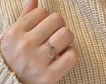 May include: A gold-colored ring featuring a double band design. One band has a rope-like texture, and the other has a delicate chain design. A small, dark blue gemstone is set on the chain band, and a whale tail charm hangs below.