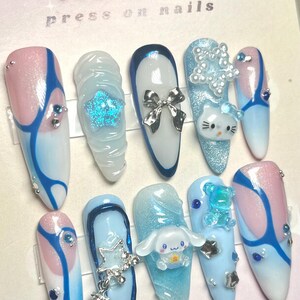 Puede incluir: Un conjunto de diez uñas postizas con temática invernal. Las uñas están decoradas con remolinos azules, blancos y rosas, purpurina y pequeños encantos. Las uñas están dispuestas en una tarjeta blanca con el texto "MONETA'S press on nails" y "@moneta_nails1".
