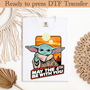 May include: White t-shirt with a graphic of a green alien character with large ears, set against an orange sunset background. The text "STAR WARS DAY MAY THE 4TH BE WITH YOU" is displayed below the image. The shirt is ready to press.