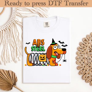 Può includere: T-shirt bianca con un disegno di un cane dei cartoni animati di Halloween. Il cane, con un cappello da strega, ha una zucca e la scritta "ABS OF STEEL". Il cane è arancione e marrone, con un colletto verde e un ragno.