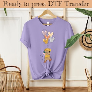 May include: A purple t-shirt with a cartoon mouse holding colorful balloons with Mickey Mouse ears. The mouse is wearing a yellow hat and holding a piece of cheese.