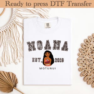 May include: White t-shirt with the word "MOANA" in a collegiate font. Below the word is an illustration of a cartoon character and the text "EST. 2016 MOTUNUI". The shirt is ready to press DTF transfer.