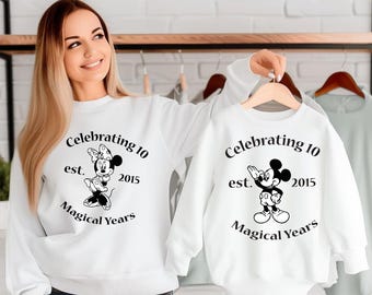 Può includere: Felpe bianche con testo nero e immagini di Topolino e Minnie Mouse. Il testo recita "Celebrating 10 est. 2015 Magical Years".