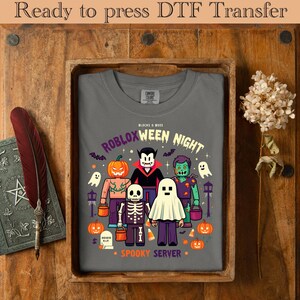 May include: A gray t-shirt with a Halloween-themed graphic featuring Roblox characters dressed as a pumpkin, vampire, zombie, skeleton, and ghost. The text reads "ROBLOXWEEN NIGHT" and "SPOOKY SERVER". The shirt is displayed in a wooden tray.