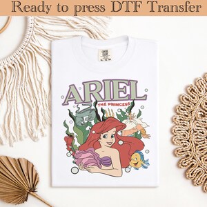 Op de afbeelding: Wit T-shirt met een kleurrijke afbeelding van Ariel uit De Kleine Zeemeermin, met de tekst "ARIEL THE PRINCESS". Het ontwerp bevat Ariel, Botje en Koning Triton, omringd door zeewier en bellen. Het shirt is klaar om te persen.