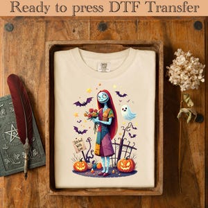 May include: A cream-colored t-shirt featuring a colorful illustration of a character holding flowers, surrounded by bats, pumpkins, and a ghost. The text "Stitched With Love" is on a small sign. The words "Ready to press DTF Transfer" are at the top.