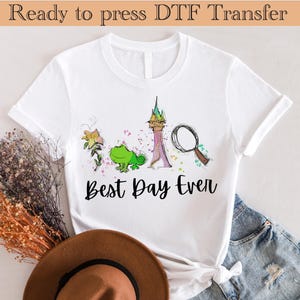May include: White t-shirt featuring a colourful graphic design of a castle, a flower, a chameleon, and a magnifying glass. The text "Best Day Ever" is printed below the graphic.