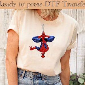 May include: A white t-shirt featuring a red and blue Spiderman graphic hanging upside down from a web. The text "Ready to press DTF Transfer" is at the top of the image.