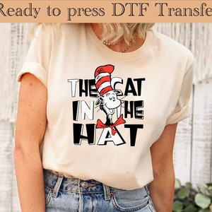 May include: A cream coloured t-shirt with a black and white graphic of the Cat in the Hat. The cat is wearing a red and white striped hat and a red bow tie.