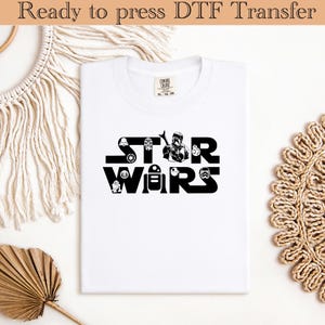 Puede incluir: Camiseta blanca con un gráfico negro de "Star Wars". El gráfico presenta varios personajes y símbolos de la franquicia Star Wars. La camiseta está doblada y lista para planchar. El texto "Ready to press DTF Transfer" está en la parte superior.