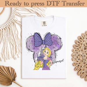 May include: White t-shirt featuring a purple Minnie Mouse-shaped design with a polka-dot bow and a Rapunzel graphic. The design includes a castle and the word "Rapunzel". The shirt is labeled "Ready to press DTF Transfer".