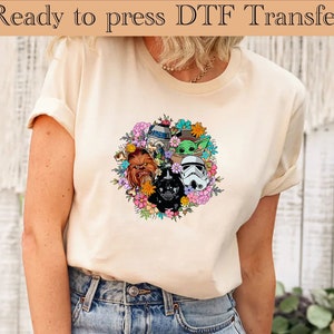 K&ouml;nnte beinhalten: Ein wei&szlig;es T-Shirt mit einem bunten Blumenmuster, das Star Wars-Charaktere zeigt, darunter Darth Vader, Chewbacca, R2-D2 und Baby Yoda.