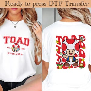 Może przedstawiać: Biała koszulka z czerwono-białą grafiką Toada z Super Mario. Przód koszulki przedstawia napis "TOAD" w czerwonych literach w stylu uniwersyteckim, z napisem "EST. 1985 SUPER MARIO" poniżej. Tył ma większą grafikę Toada.