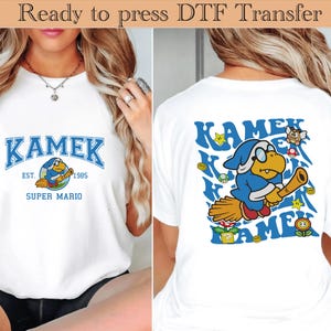 May include: White t-shirt featuring a blue and white graphic of Kamek from Super Mario. The front design includes the word "KAMEK" in blue, with "EST. 1985 SUPER MARIO" below. The back design shows Kamek on a broom with stars and coins.