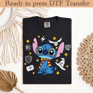 Puede incluir: Camiseta negra con un personaje de dibujos animados azul con gafas y bufanda, rodeado de estrellas amarillas y gráficos temáticos de Harry Potter. El texto "Ready to press DTF Transfer" está en la parte superior.