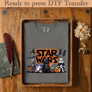 May include: A gray t-shirt featuring a Star Wars graphic with characters like Darth Vader, Yoda, and a Stormtrooper. The design includes the text "STAR WARS" in orange. The shirt is displayed in a wooden tray.