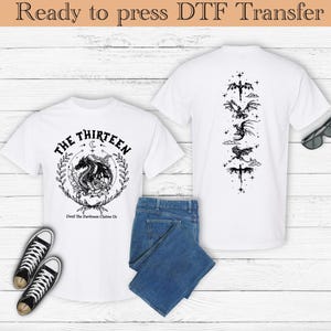 以下が含まれることがあります： 黒いグラフィックデザインが施された白いTシャツ2枚。前面のシャツには、「The Thirteen」と「Until the Darkness Claims Us」というテキストとドラゴンが描かれています。背面のシャツには、星と三日月が描かれた5匹のドラゴンが縦に並んでいます。