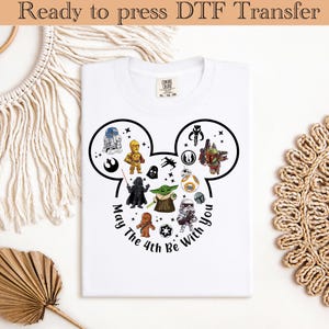 Puede incluir: Camiseta blanca con un diseño negro en forma de Mickey Mouse lleno de personajes de Star Wars. El texto "May The 4th Be With You" está impreso alrededor del diseño. Transferencia DTF lista para planchar.