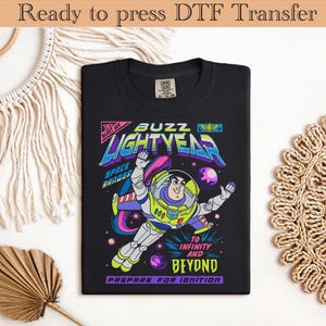 May include: Black t-shirt featuring a colorful graphic of Buzz Lightyear in a space scene. The design includes the text "BUZZ LIGHTYEAR", "TO INFINITY AND BEYOND", and "PREPARE FOR IGNITION". The shirt is ready for DTF transfer.