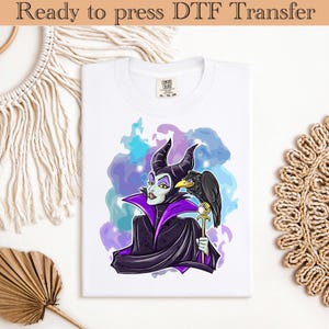 May include: White t-shirt featuring a graphic of a character with black horns, a black raven, and a staff. The character is wearing a purple and black outfit. The background has blue and purple watercolor-style splashes. Text at the top reads "Ready to press DTF Transfer."
