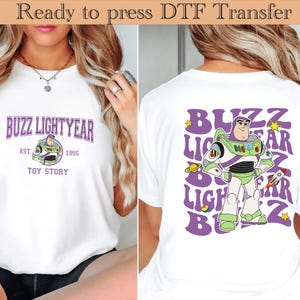 以下が含まれることがあります： 白いTシャツ。前面には紫色の「Buzz Lightyear」の文字、バズの画像、そして「Toy Story」の文字がデザインされています。背面には、より大きなバズのグラフィックがあります。