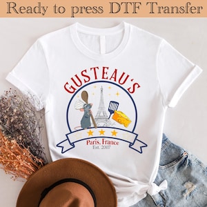 May include: White t-shirt with a graphic of Remy the rat from the movie Ratatouille. The graphic includes the text "Gusteau's" in red and blue, a drawing of the Eiffel Tower, a spatula, and a piece of cheese. The text "Paris, France Est. 2007" is also included in the graphic.