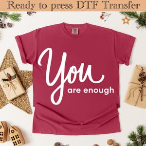 May include: A maroon t-shirt with the words "You are enough" in a white script font. The shirt is displayed on a white surface with festive decorations, including wrapped gifts and greenery.