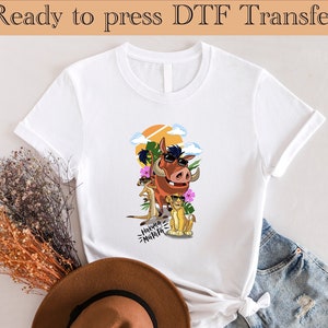 Op de afbeelding: Wit T-shirt met een cartoon afbeelding van Pumbaa uit The Lion King met een zonnebril op een bloemige achtergrond. De tekst "Hakuna Matata" is onder de afbeelding gedrukt.