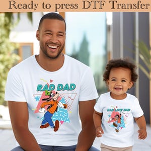 Puede incluir: Camisetas blancas con gráficos a juego. Una camiseta dice "RAD DAD" con un personaje de dibujos animados con chaqueta naranja, pantalones azules y sombrero verde. La otra camiseta dice "RAD LIKE DAD" con un personaje de dibujos animados similar.