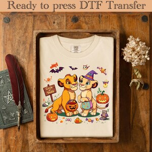 May include: Cream-colored t-shirt featuring a Halloween-themed graphic with two lion cubs, pumpkins, bats, and the text "Circle of Treats". The shirt is displayed in a wooden tray on a wooden surface.